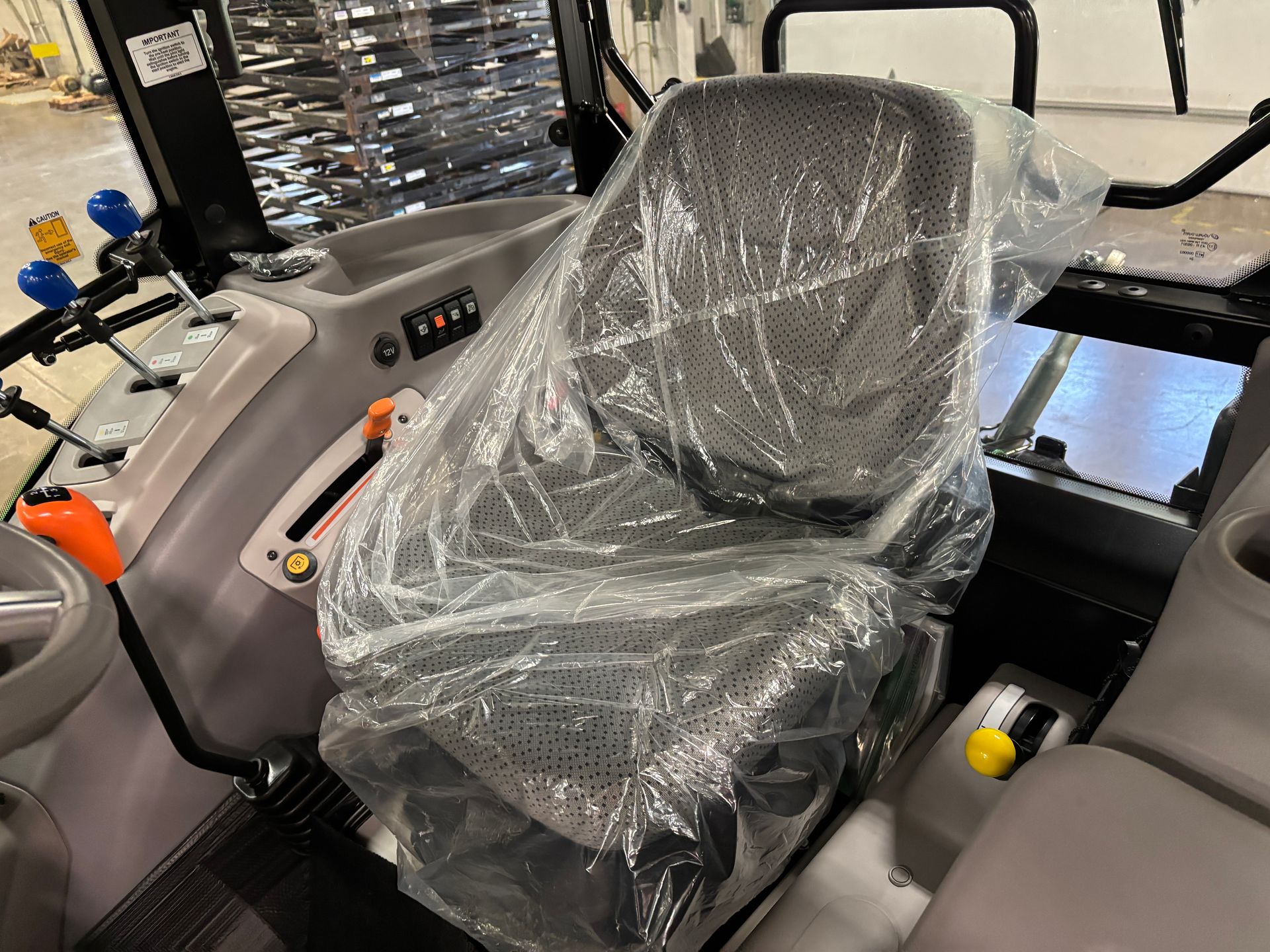 Tractor interior with seat covered in plastic. Levers, controls, and steering wheel are visible. Gray and black tones.