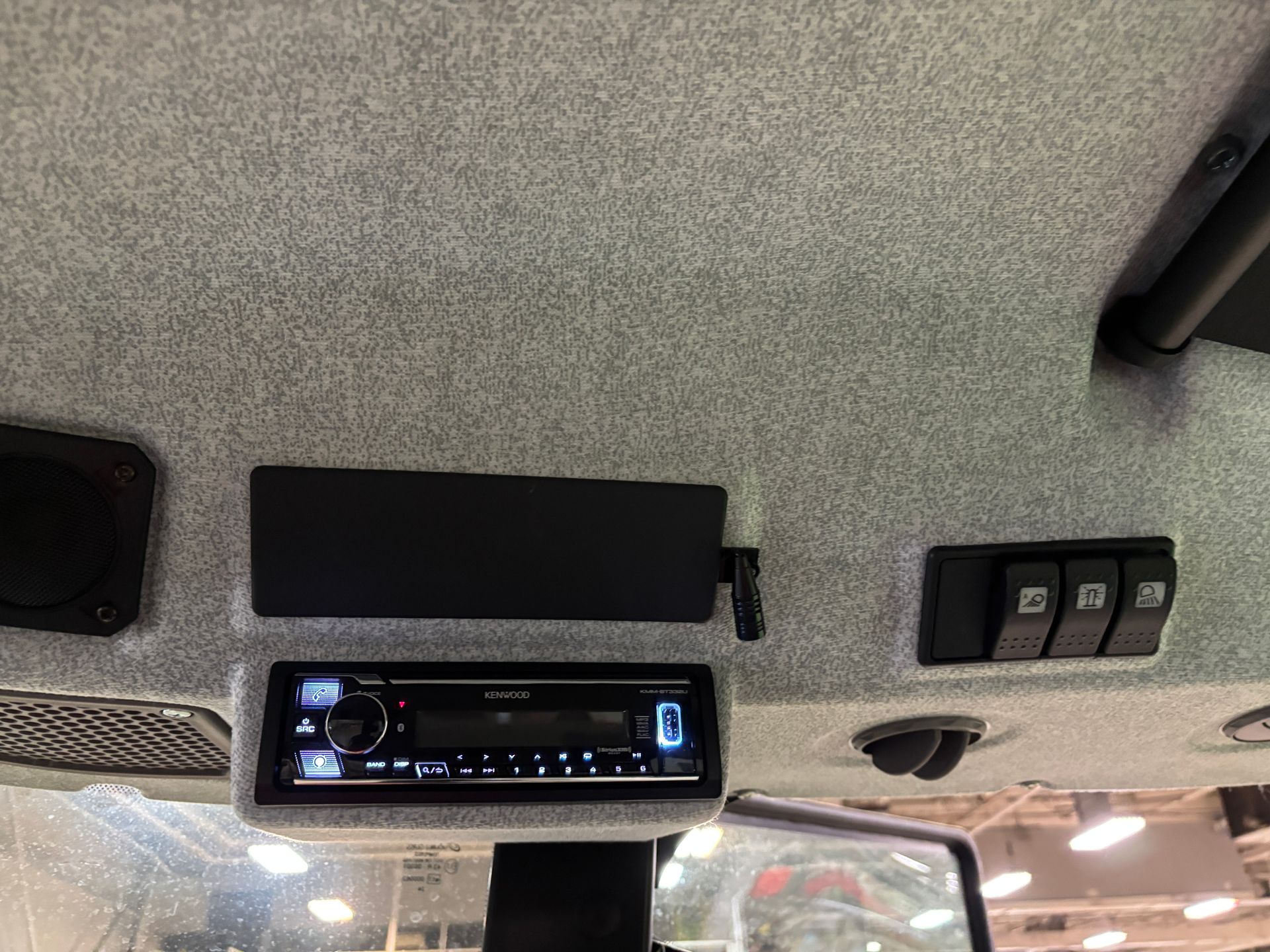 Interior view of a vehicle's ceiling: radio, switches, and speakers against a grey fabric backdrop.