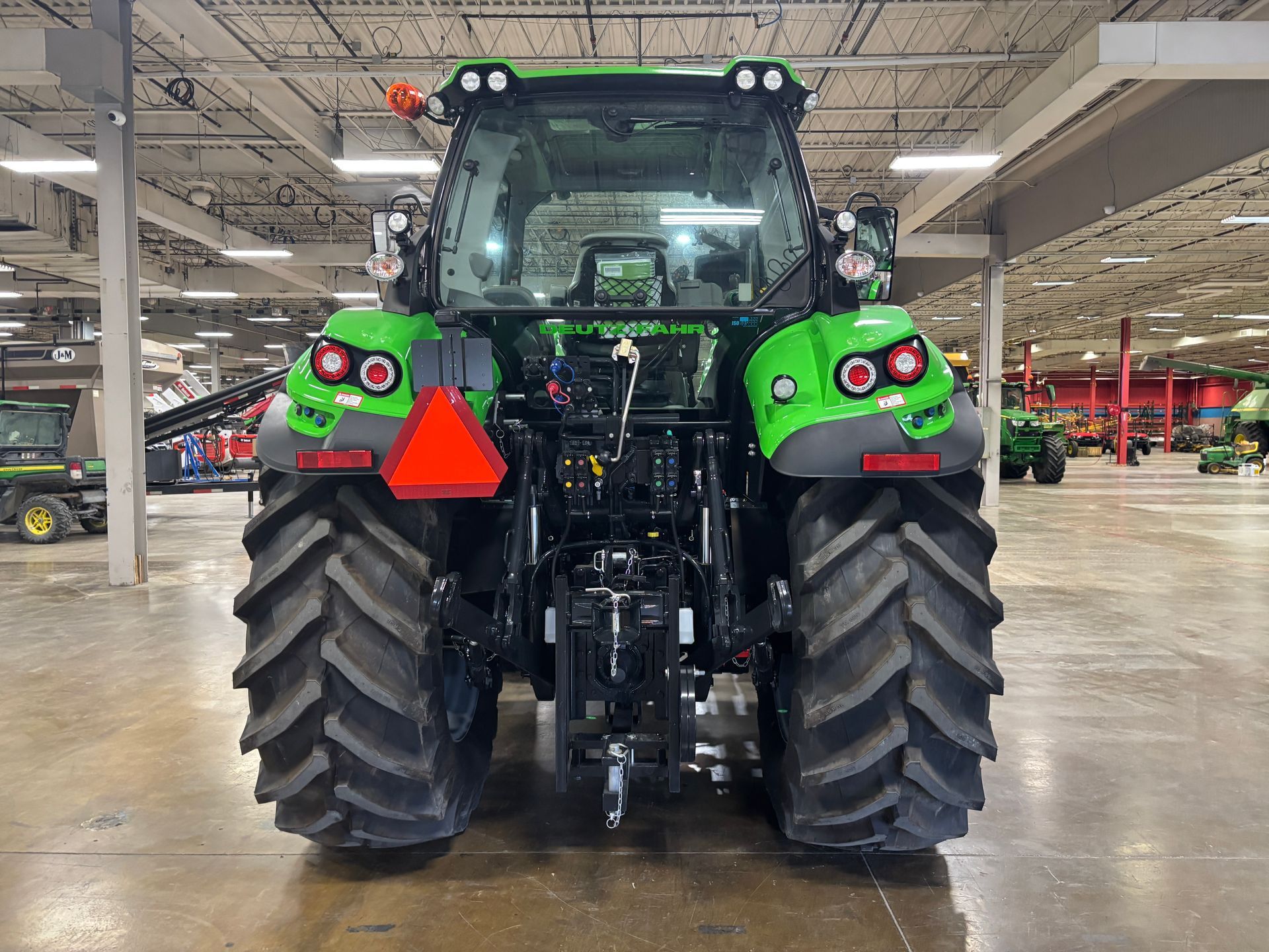 Rear view of a green tractor with large tires, in a warehouse setting, with red triangle safety sign.