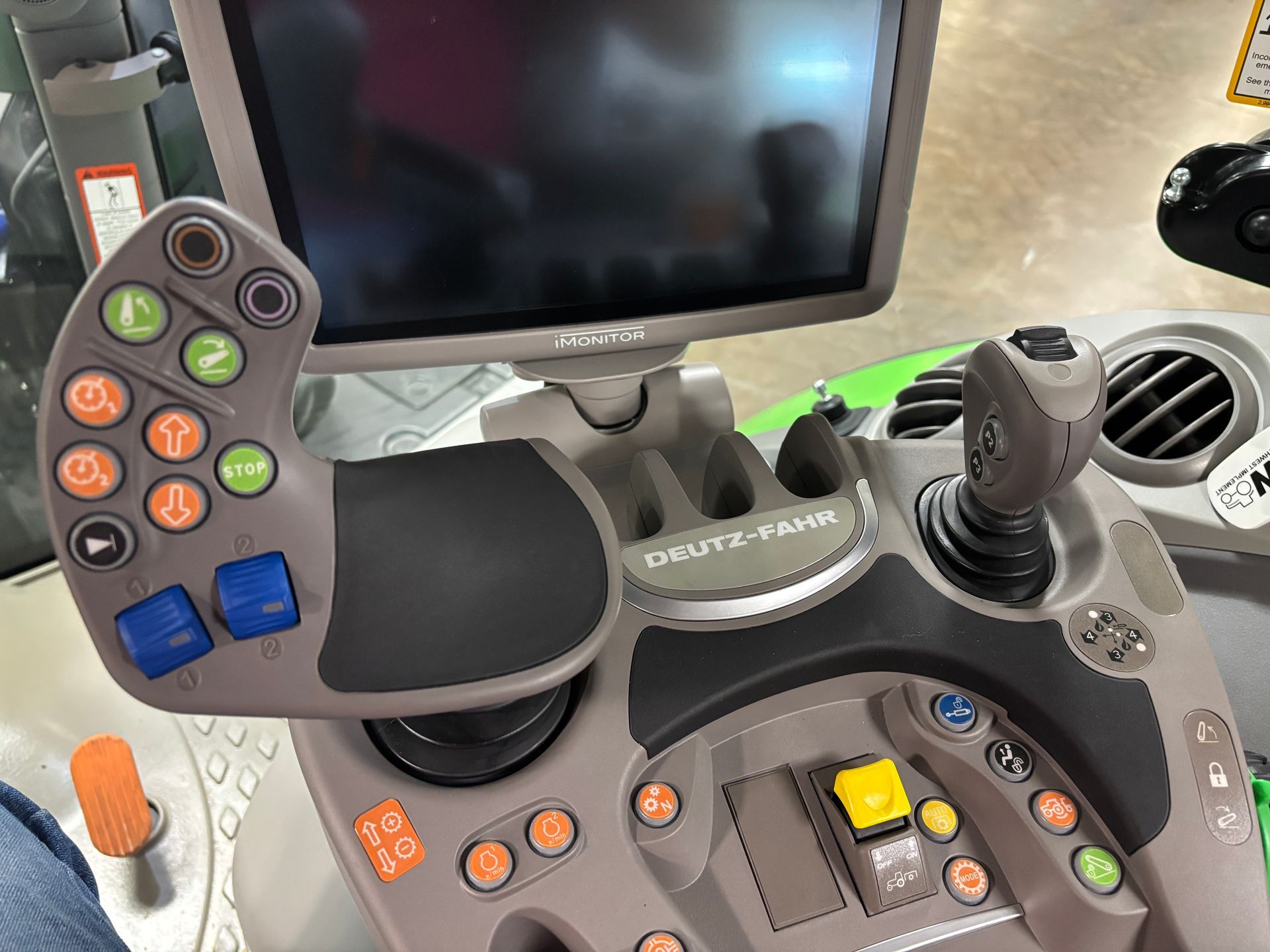 Interior view of a tractor console with a steering wheel and joystick, various orange and blue buttons, and a digital display screen.