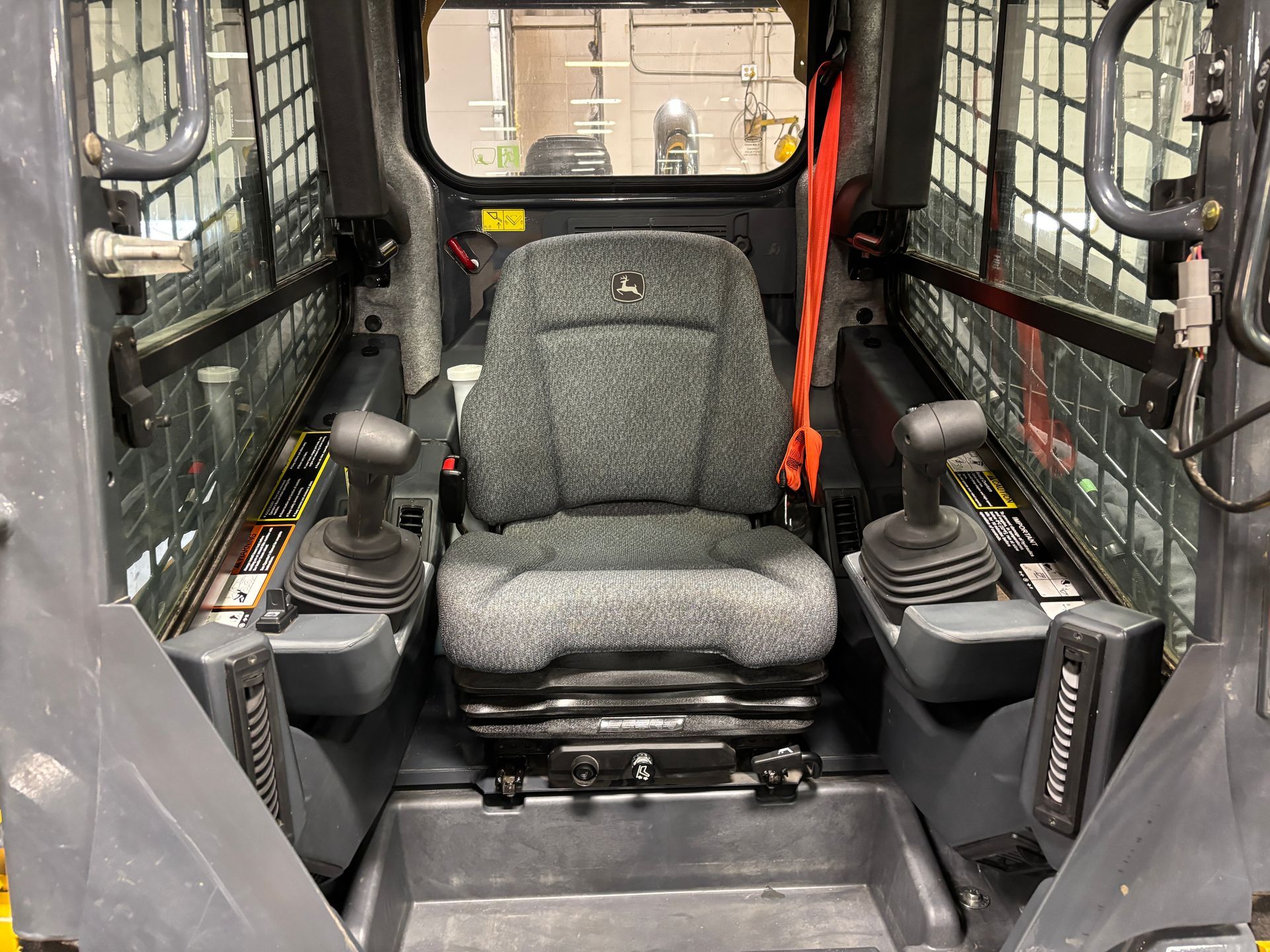 Interior of a compact track loader cab with gray seat, joysticks, and safety features.