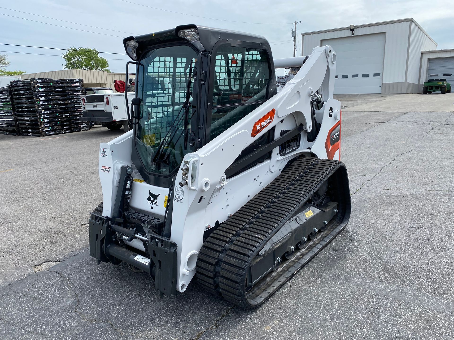 White Bobcat compact track loader on asphalt, tracks visible. Industrial setting with warehouse and sky.
