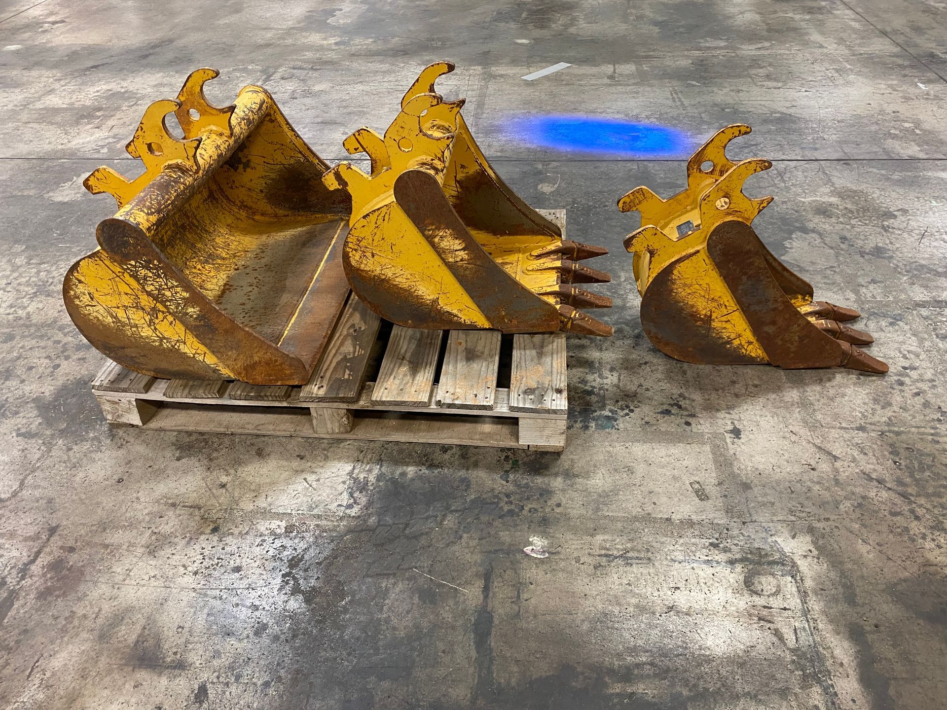 Yellow excavator buckets sit on a wooden pallet on a concrete floor.