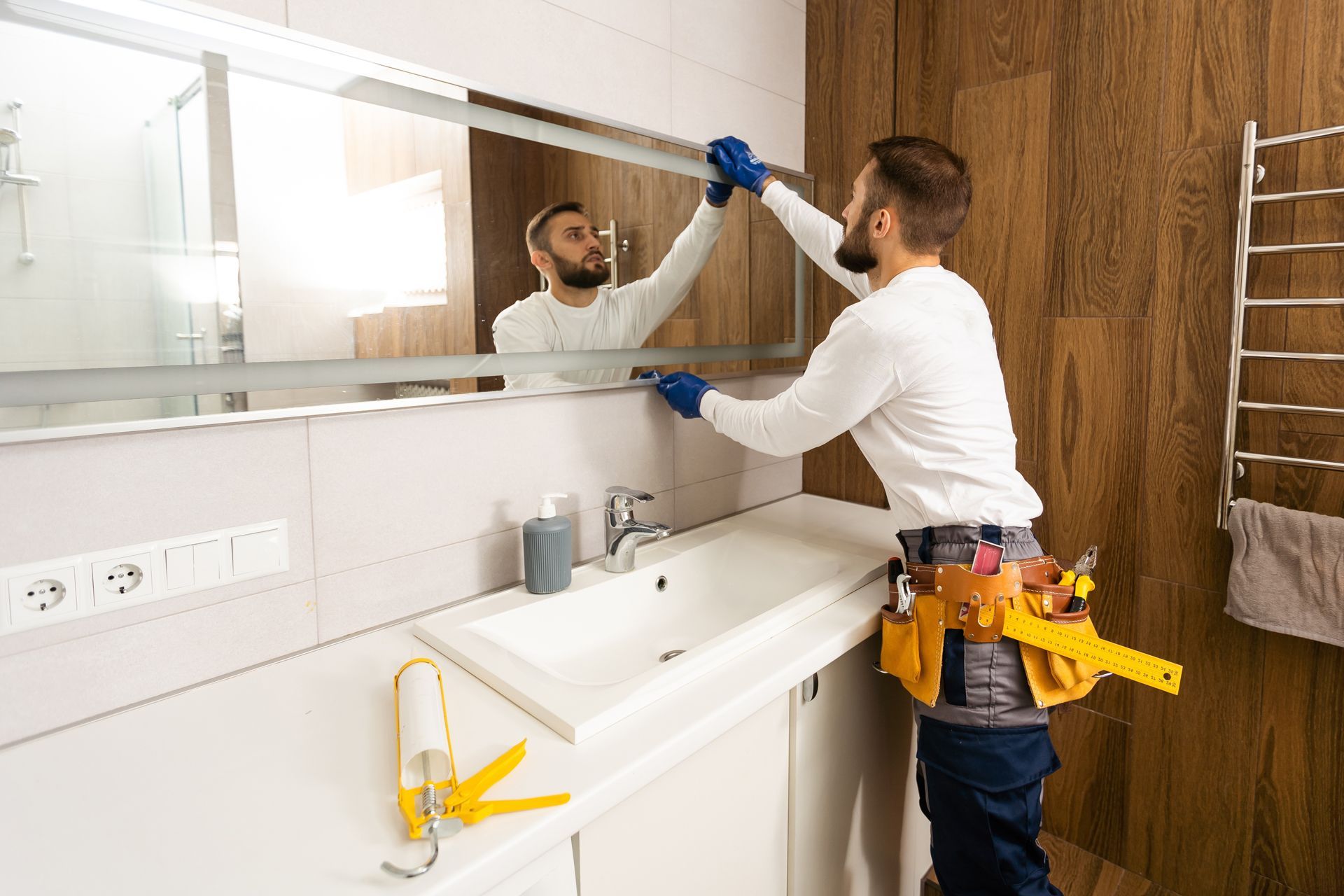 Worker installing a bathroom mirror above a sink, with tools on a utility belt.