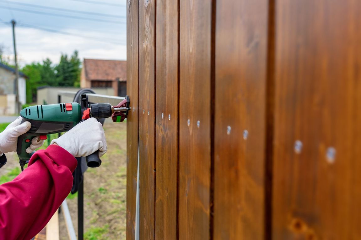 Gloved hands using a power drill to install screws into a wooden fence panel outdoors