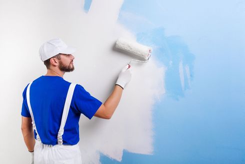 Painter rolling white paint on a wall, covering blue paint in a bright room.