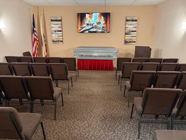 A funeral viewing room with rows of chairs facing a casket centered on a red-skirted table beneath a fireplace monitor.