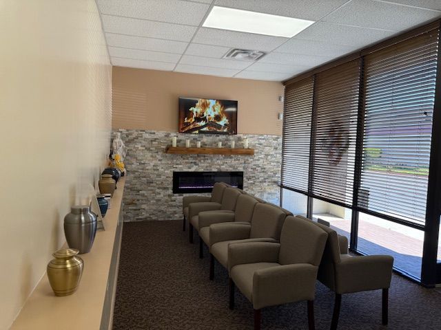 A waiting room with chairs facing a fireplace and a wall-mounted TV, with urns displayed on a shelf in the foreground.