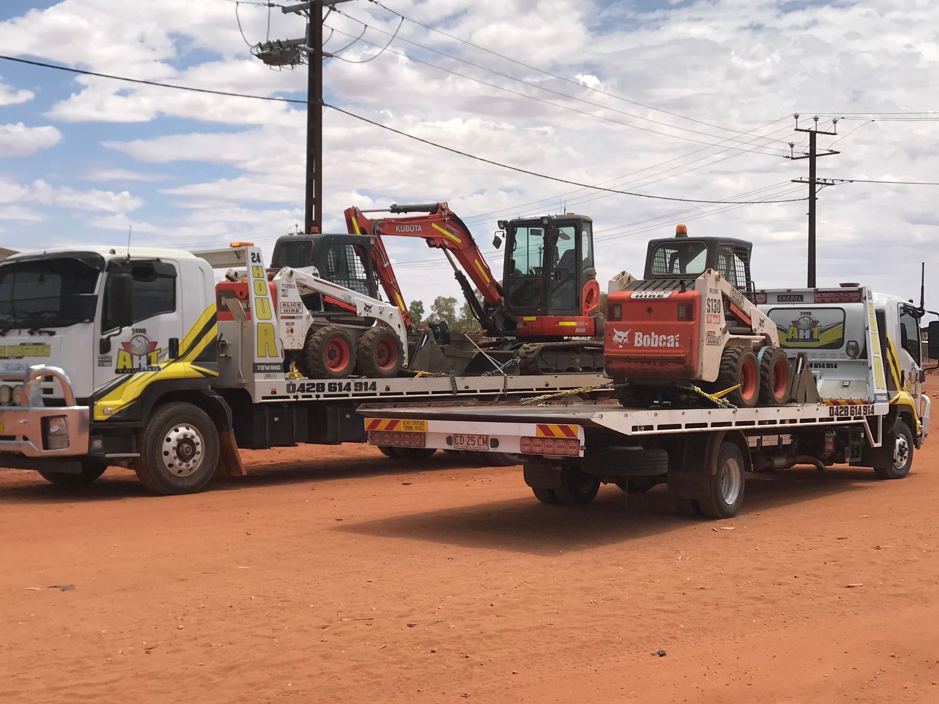 24-hour tilt tray towing service with AHT trucks carrying machinery in a rural construction setting.