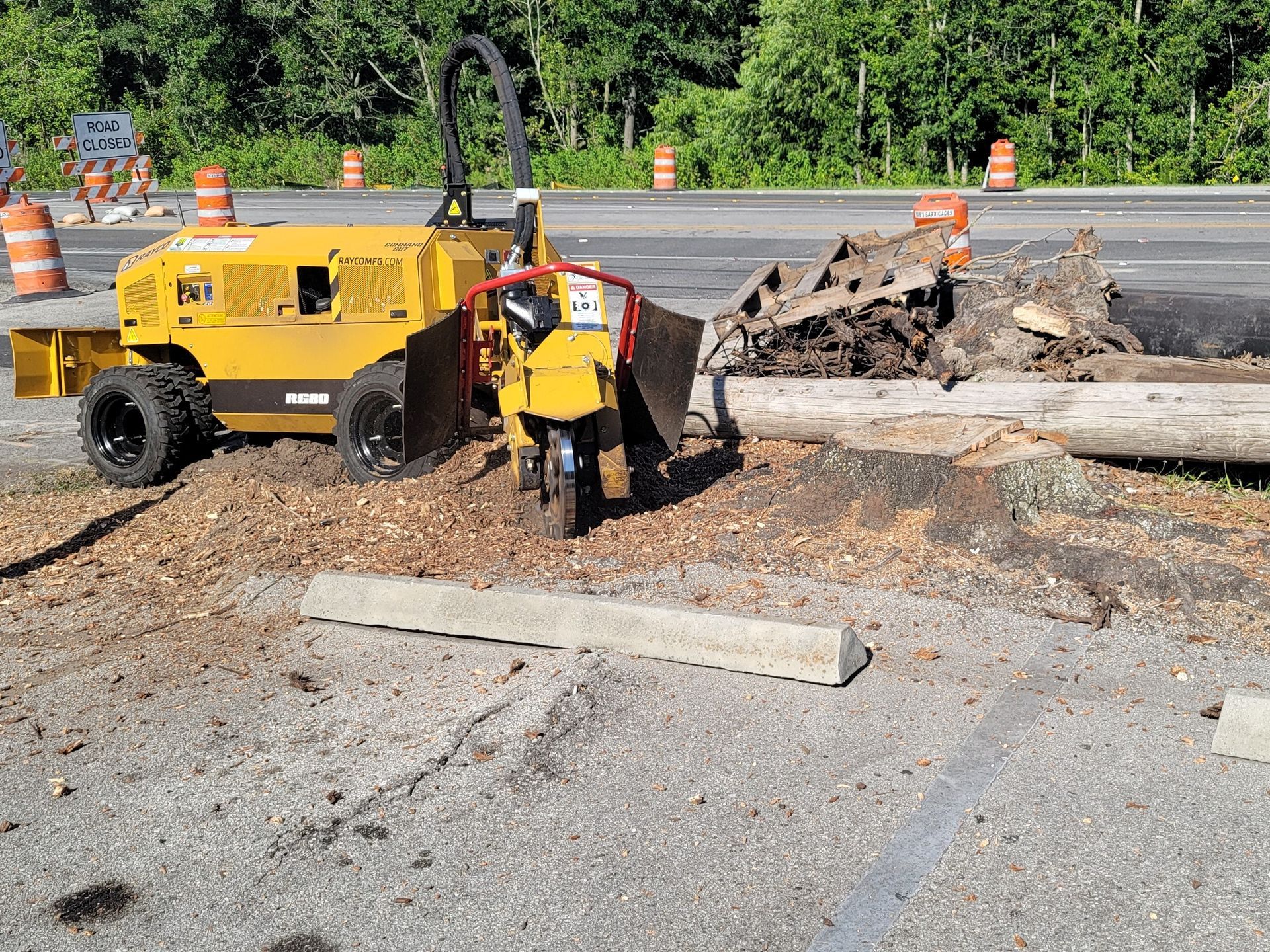 A yellow tractor is cutting a tree stump on the side of the road.