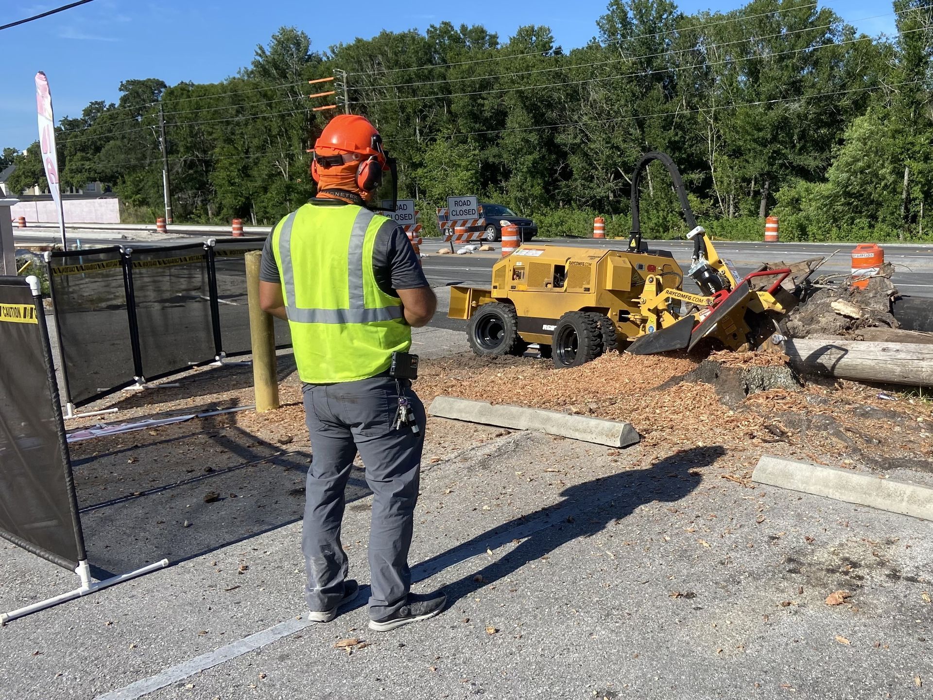 A construction worker is standing next to a stump grinder in a parking lot.