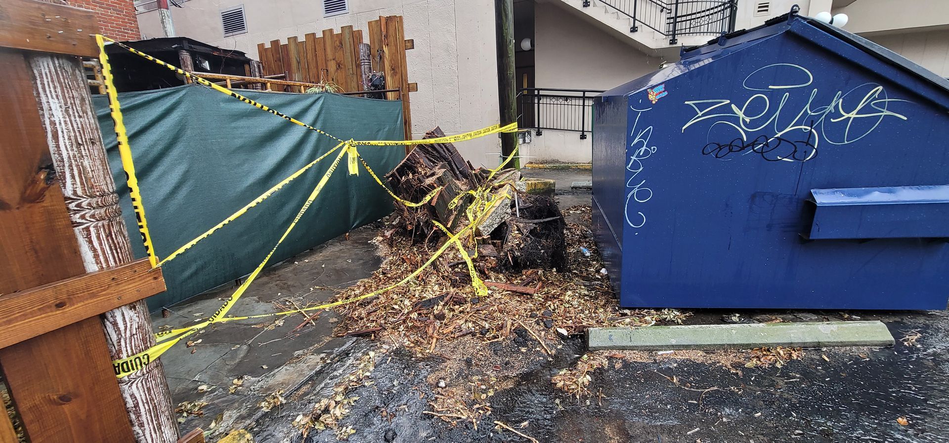 A blue dumpster with graffiti on it is surrounded by yellow tape.