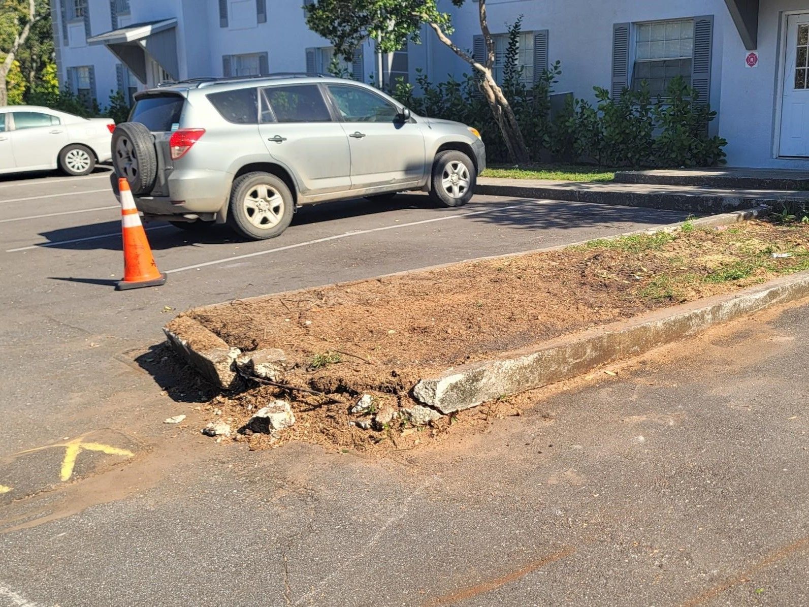 A silver suv is parked in a parking lot next to an orange cone by a broken curb.