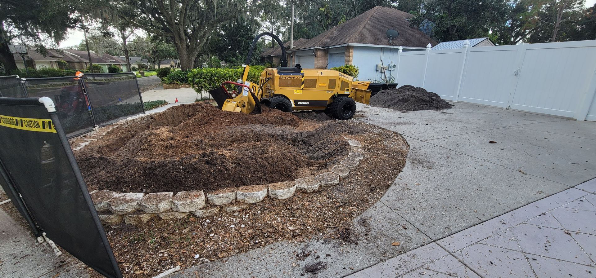 A yellow tractor is cutting a tree stump in a driveway.