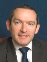 Man in a suit smiles, dark blue tie, against a blue background.