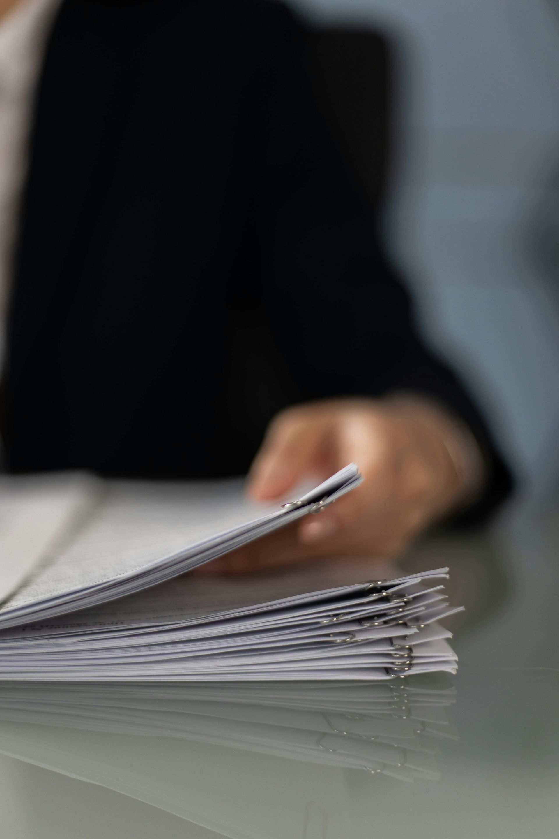 Person in black suit reviewing a stack of papers on a glass table.
