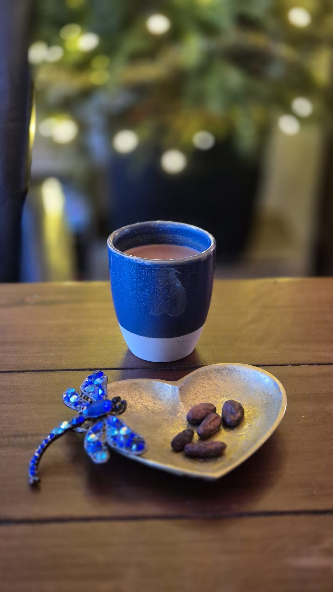 Een blauw-witte mok cacao op een houten tafel, met een hartvormig schaaltje cacaobonen en een blauwe libel als decoratie.