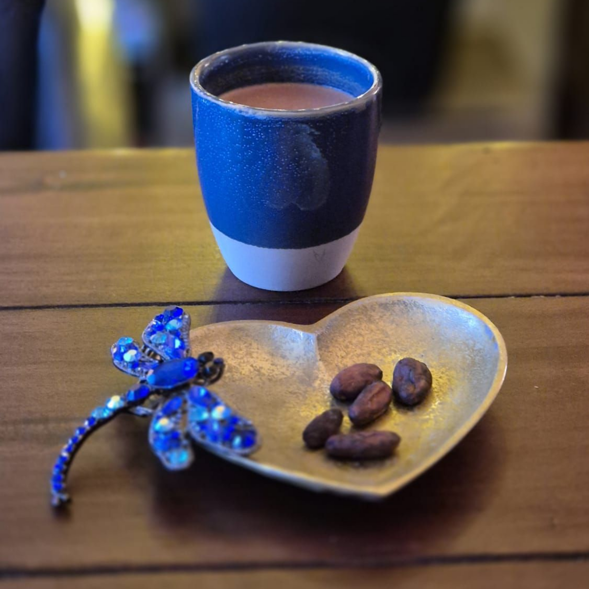 Een blauw gespikkelde mok met warme chocolademelk en een hartvormig schaaltje cacaobonen, naast een blauwe libelle