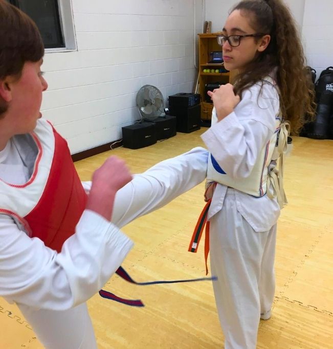 Family in Jiu-Jitsu gis poses. Indoors, blue mats, a smiling woman in the middle, around a seated man with a beard.