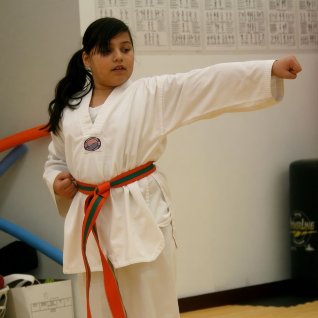 Two children practicing Brazilian Jiu-Jitsu on a mat. One child in white gi has the other in a leg lock. A girl watches.