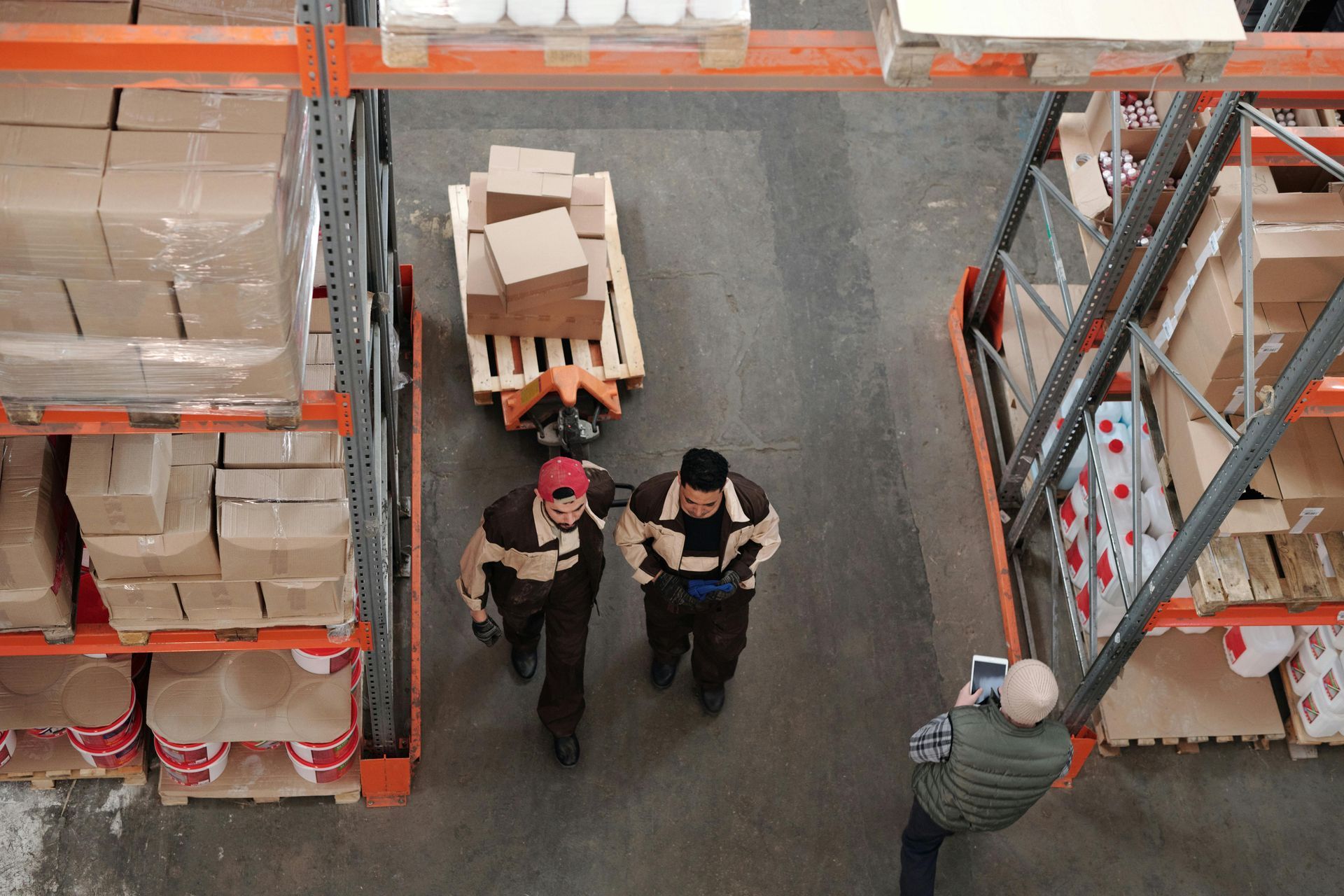 Workers in beige uniforms walk between warehouse shelves, one pulling a pallet of cardboard boxes while another observes.