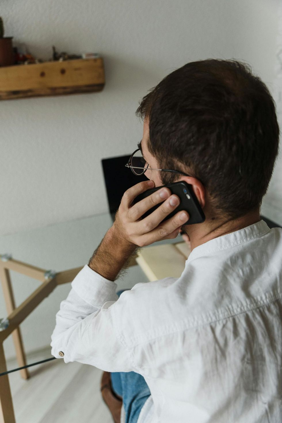 A person in a white button-down shirt sitting at a desk, talking on a smartphone with a laptop in the background.