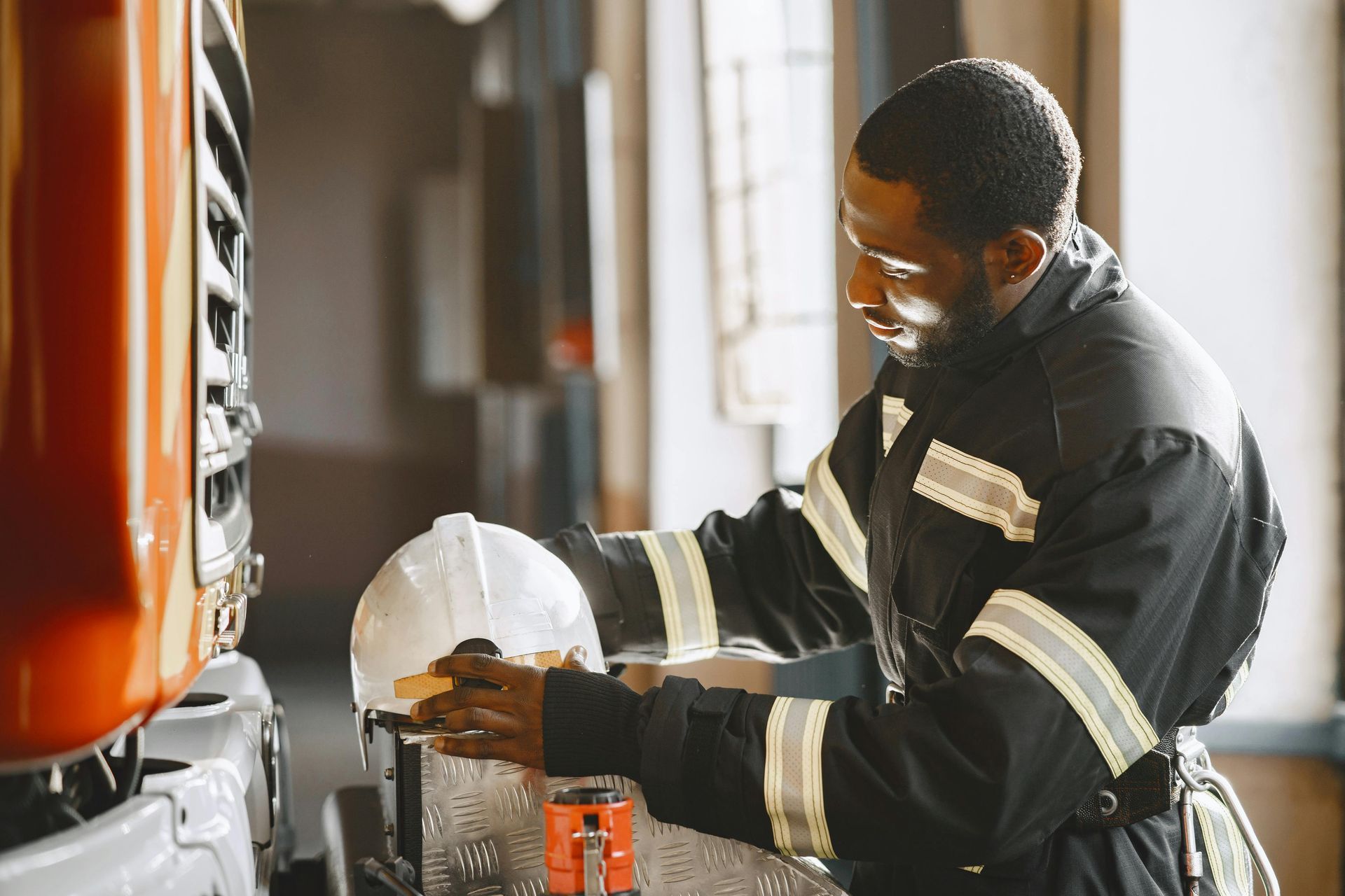 A worker in a high-visibility uniform inspects a white hard hat near a large orange truck in a garage.