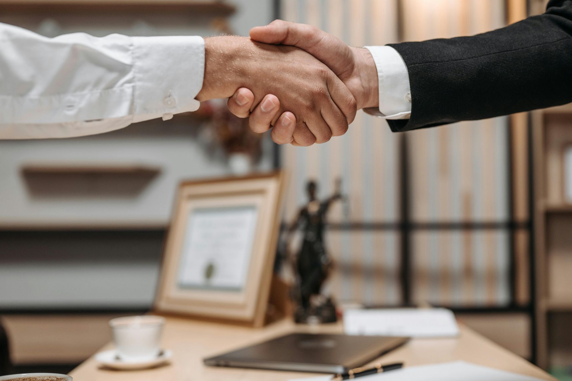 Two people in professional attire shake hands across a desk in a law office, with a legal statue visible in the background.