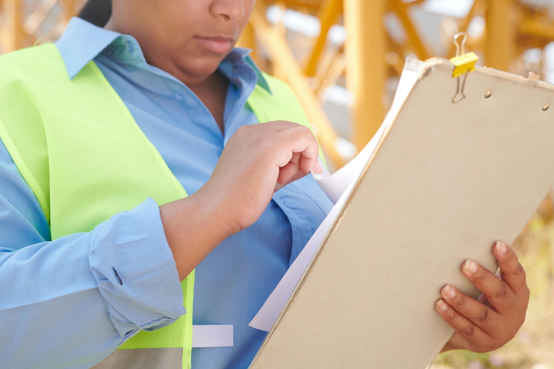 A person wearing a blue shirt and neon yellow safety vest reviews papers attached to a clipboard in an outdoor setting.