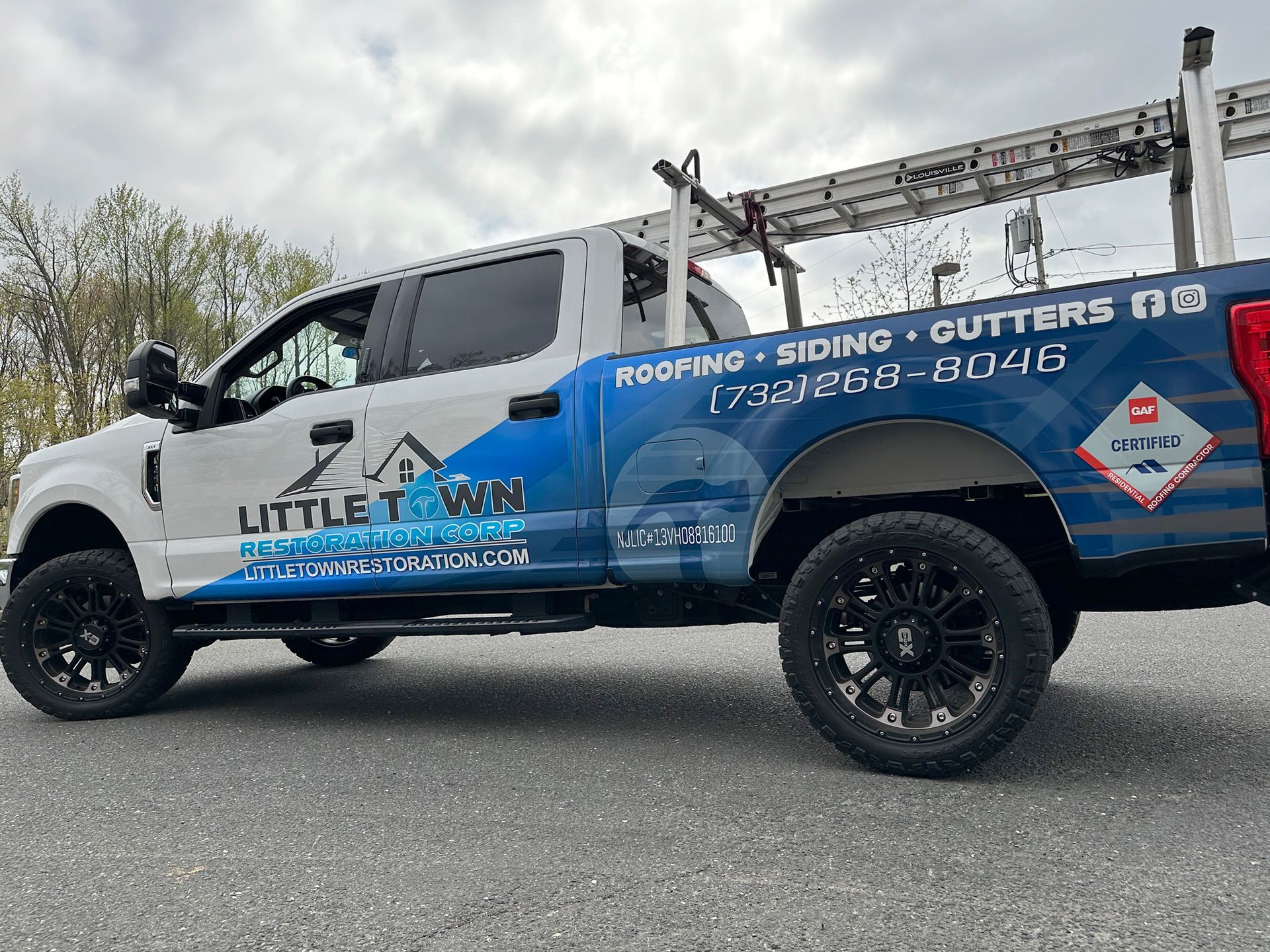 A white and blue truck with a ladder on the back is parked on the side of the road.