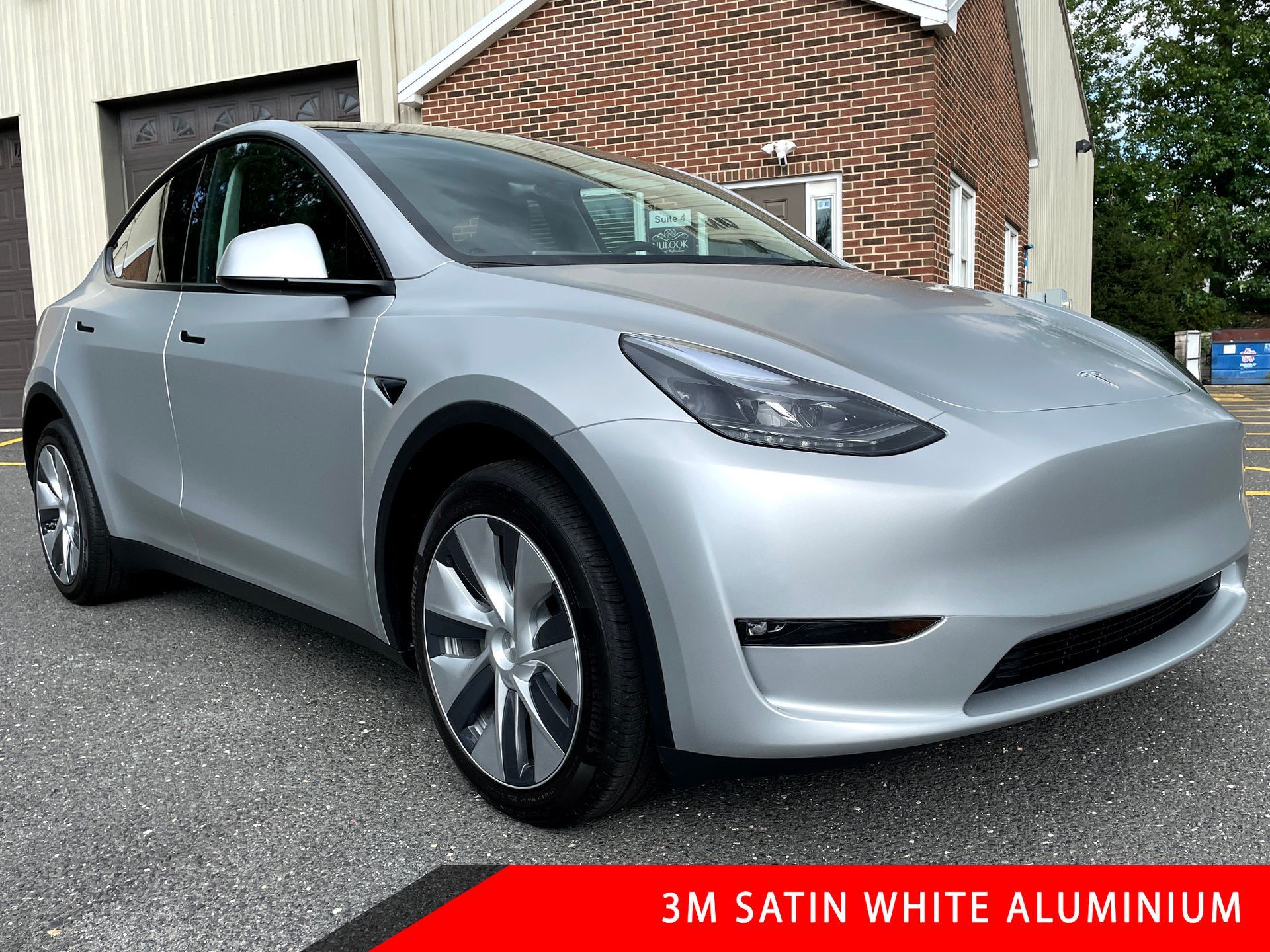 A silver tesla model y is parked in front of a brick building.