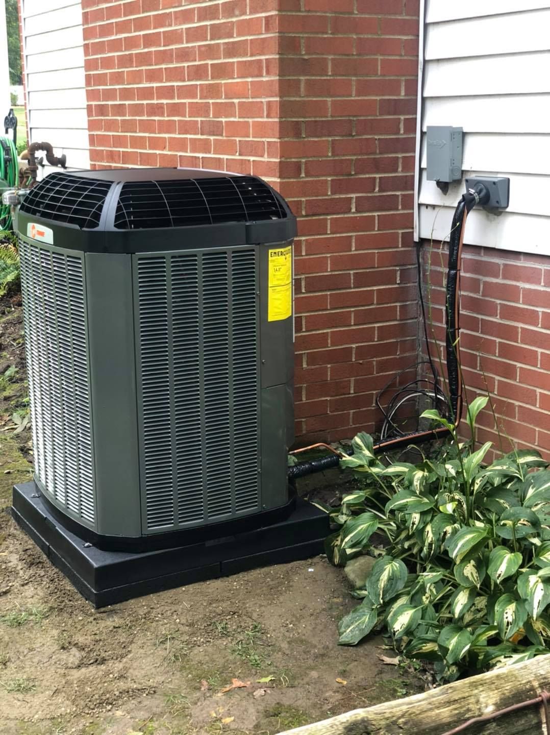 Air conditioner unit on a black base next to brick wall and bushes.