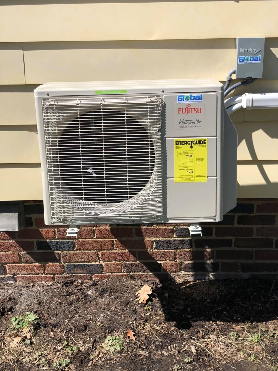 Fujitsu AC unit mounted on a brick wall, beige siding background, power lines, and a yellow energy label.