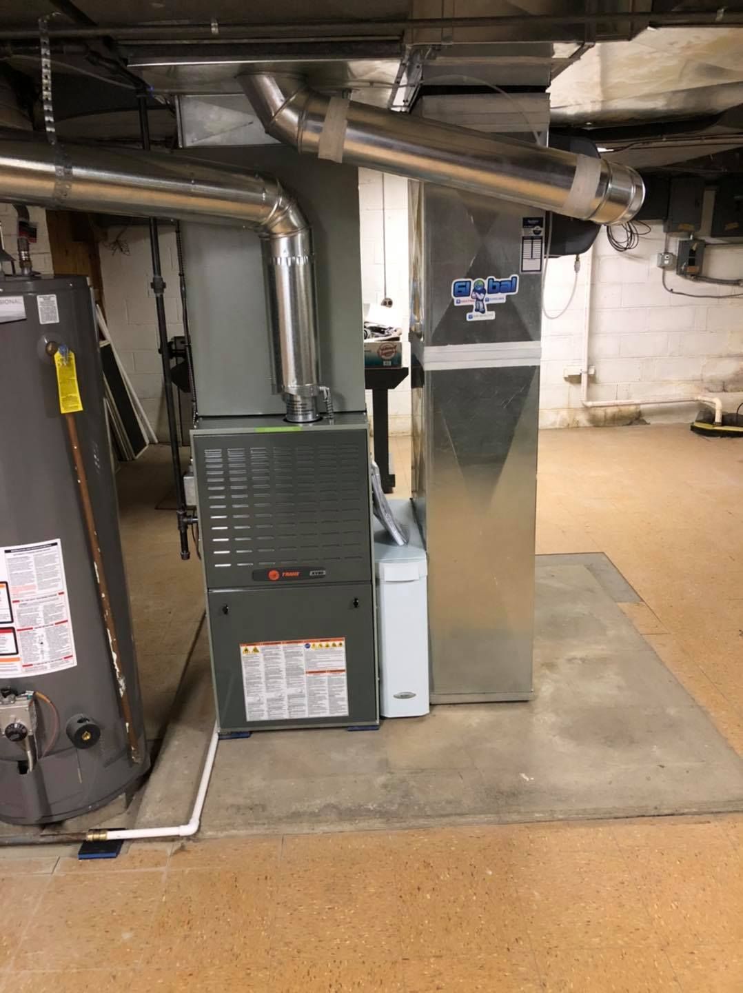 Furnace and water heater in a basement. Metal ducts and pipes are overhead. Gray and white appliances on a concrete pad.