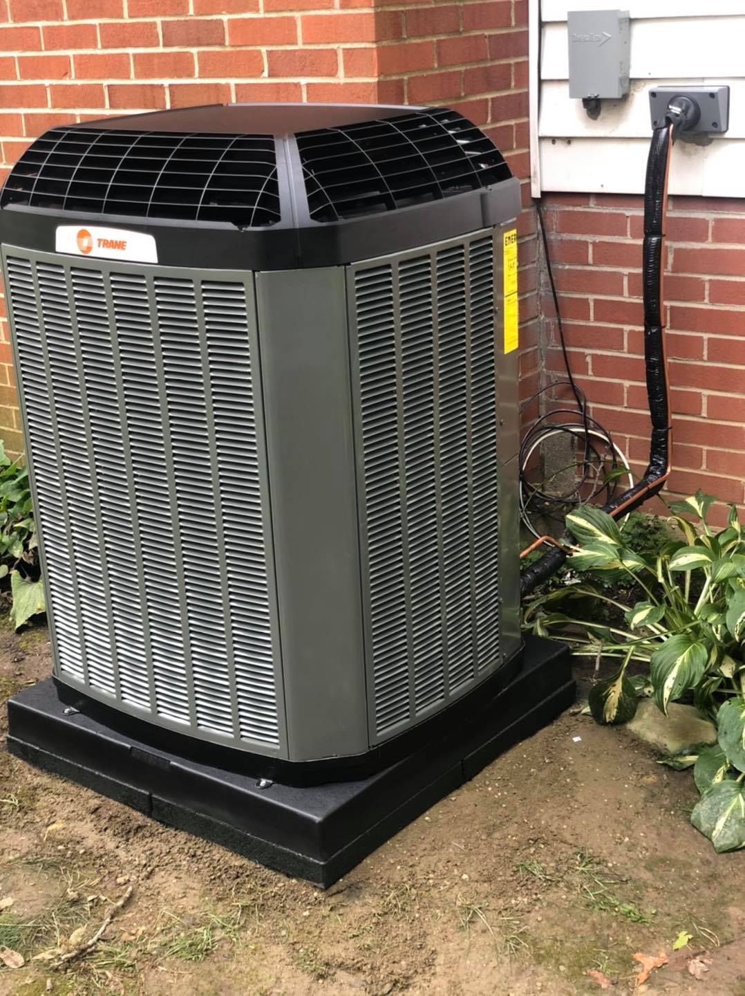 Gray air conditioning unit on a black platform near a brick wall and plants.
