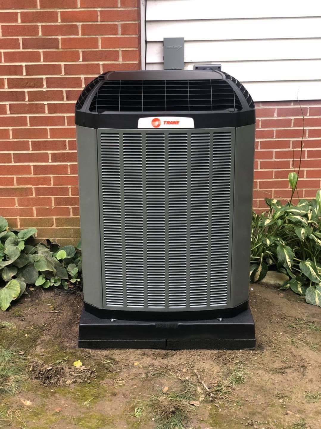 Gray Trane air conditioner unit next to a brick wall and vegetation.