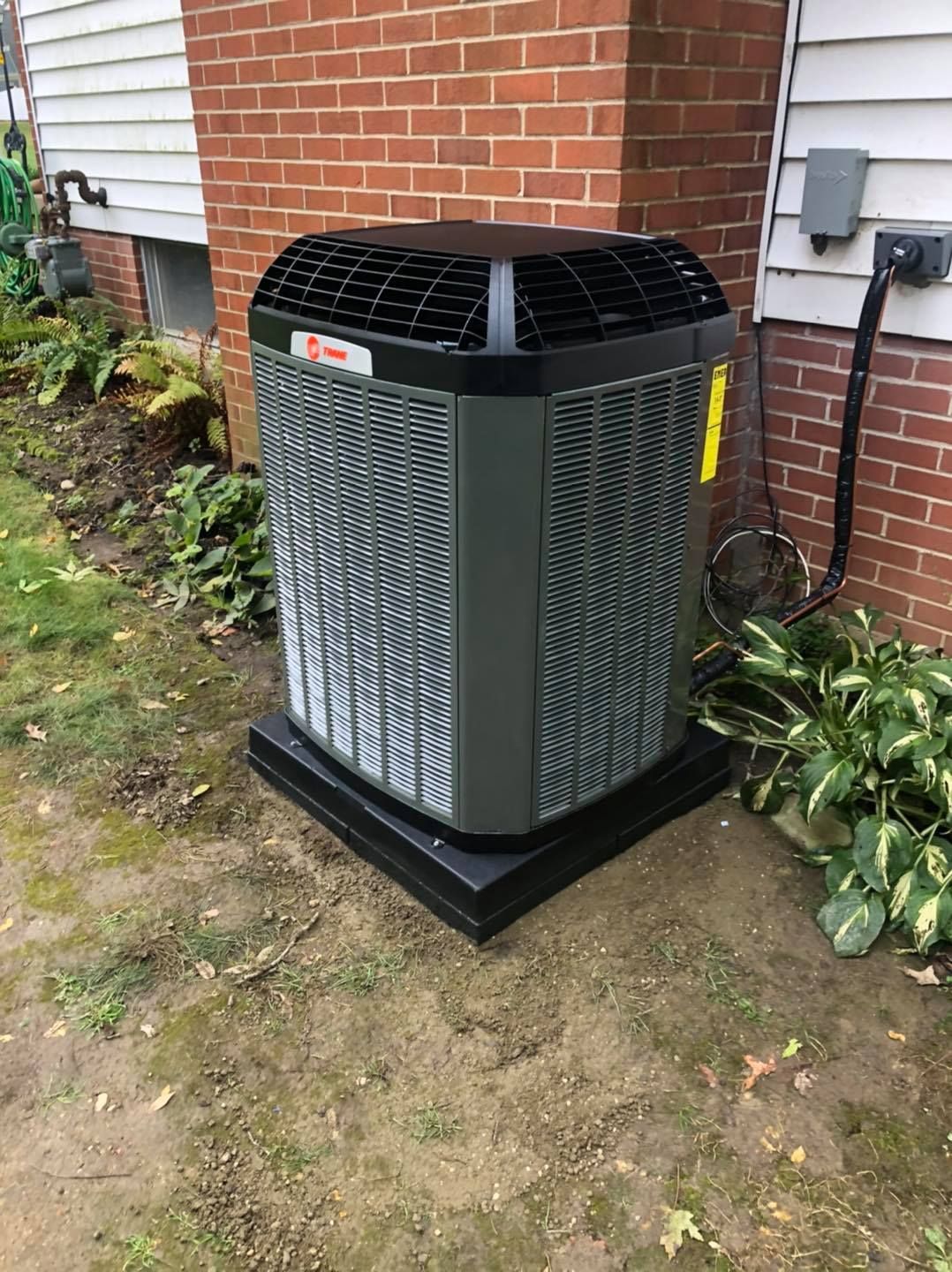 An outdoor air conditioning unit on a black base, next to a brick wall and foliage.