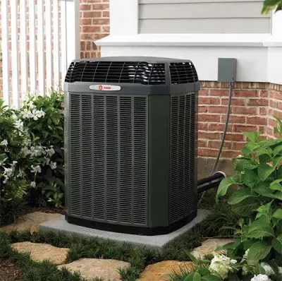 Dark gray air conditioning unit outside a brick building, sitting on a stone slab in a landscaped area.