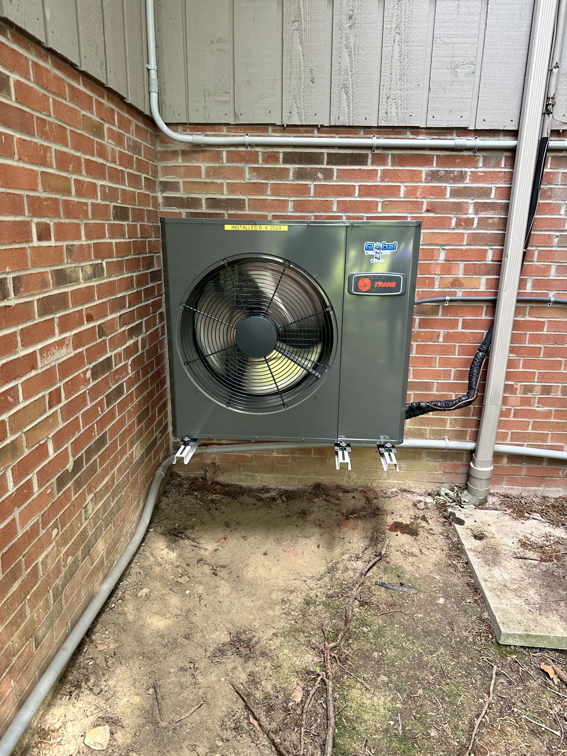 Outdoor HVAC unit mounted on brick wall, with fan facing out. Grey, with silver pipes.