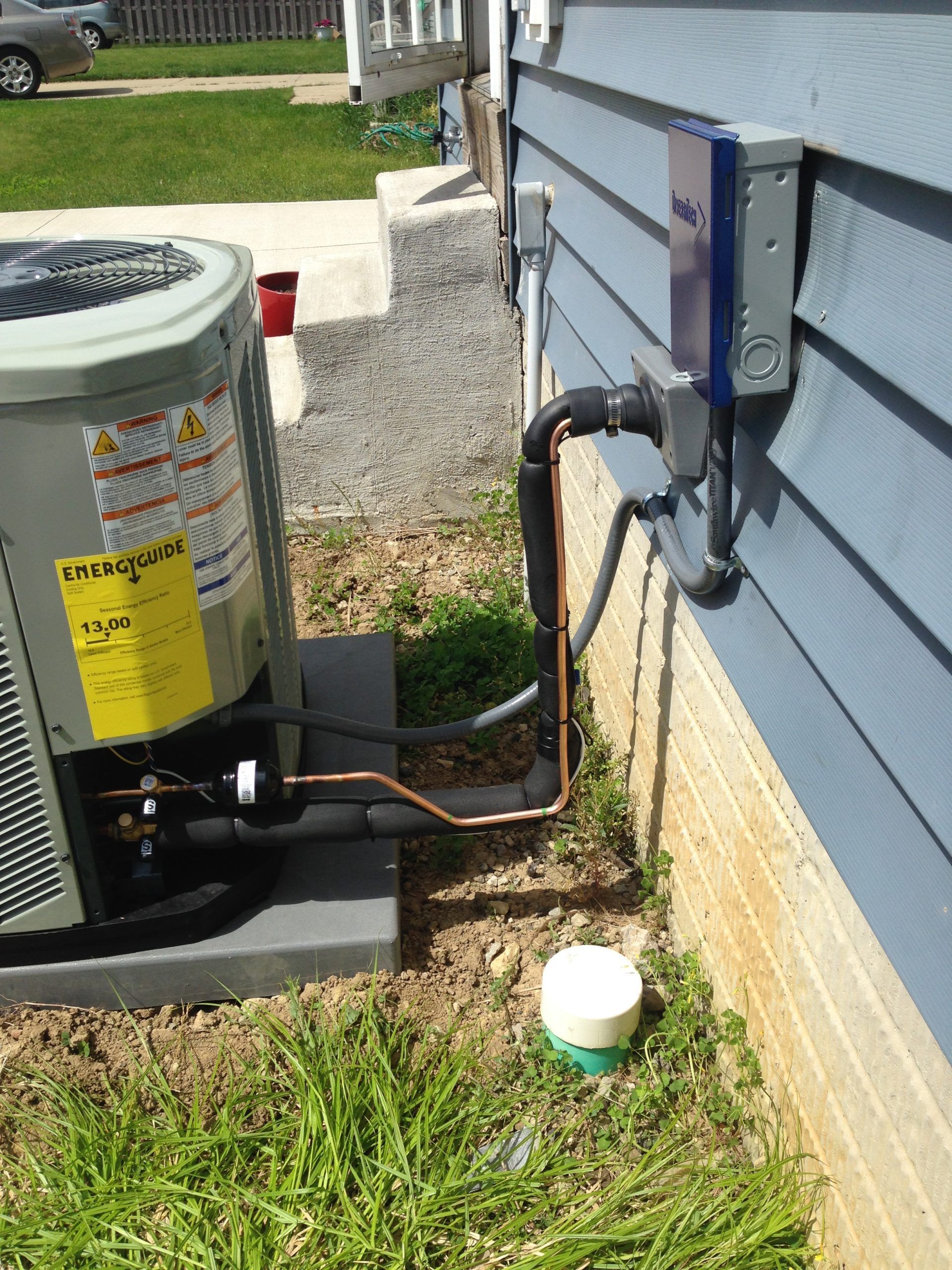 HVAC unit outside blue house, with copper pipes and electrical box. Green grass and gravel path.
