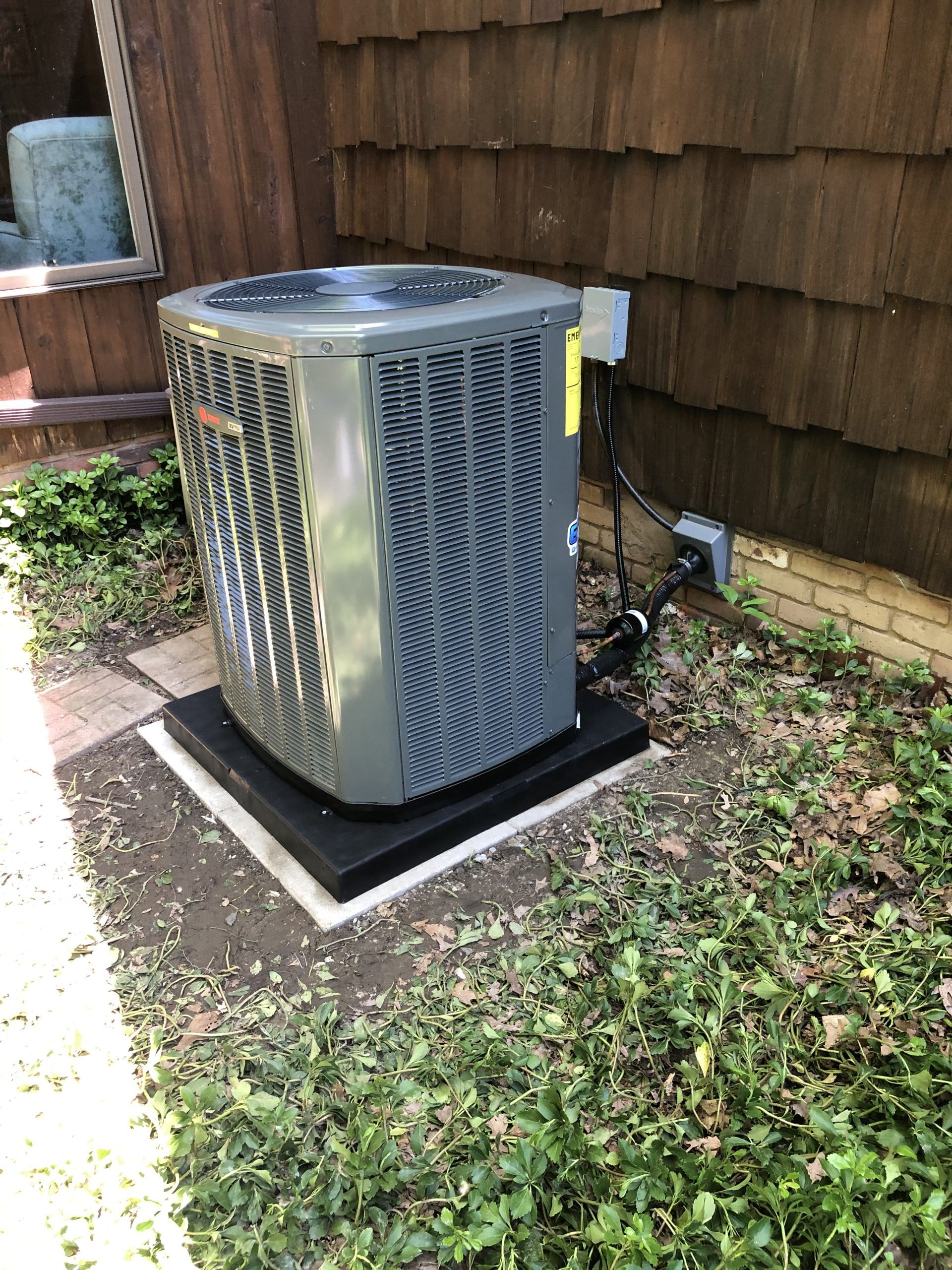 An outdoor air conditioning unit sits on a black platform near a building with brown siding.