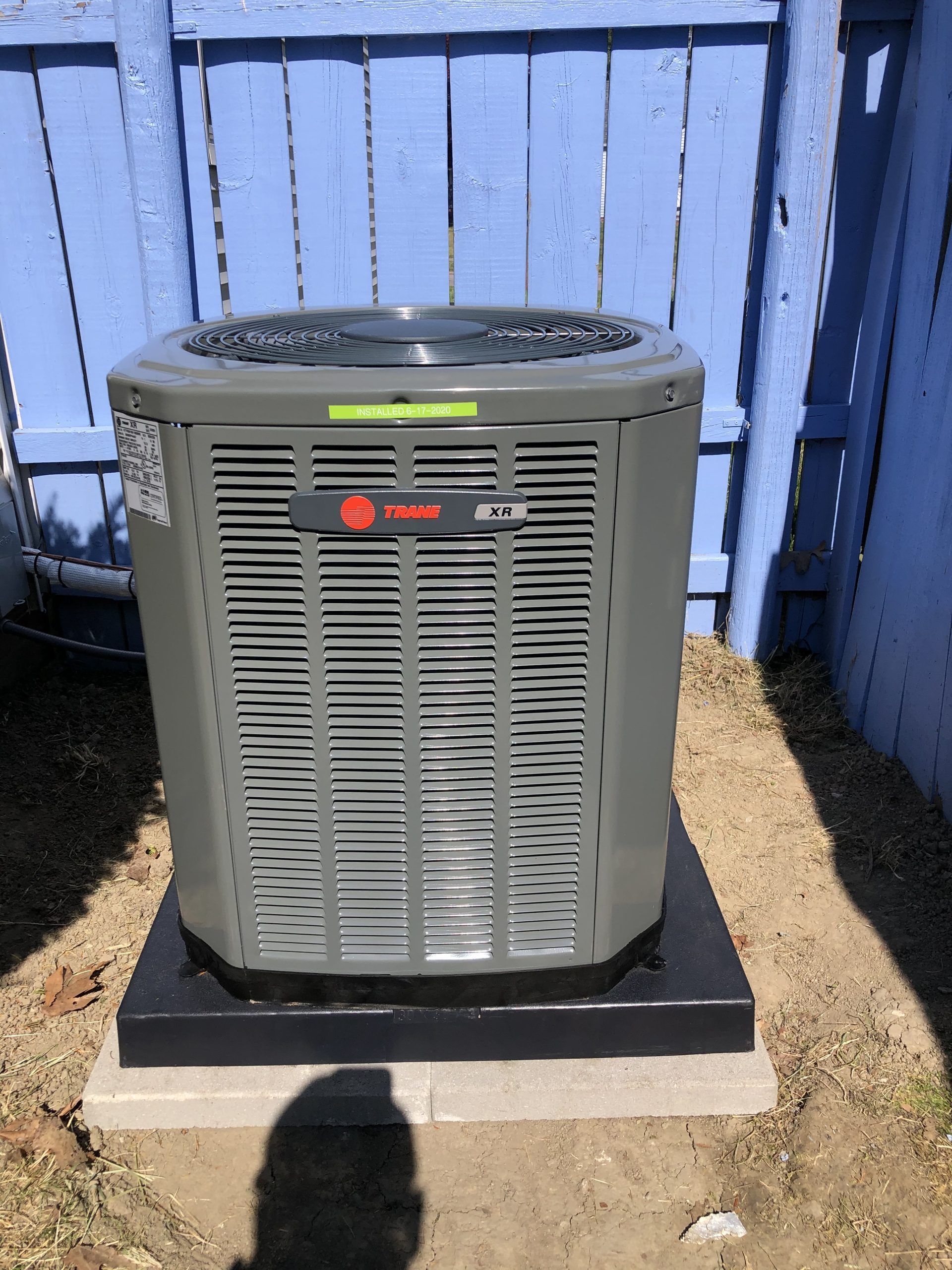 Gray Trane air conditioner unit, set on a black base, against a blue wooden fence.