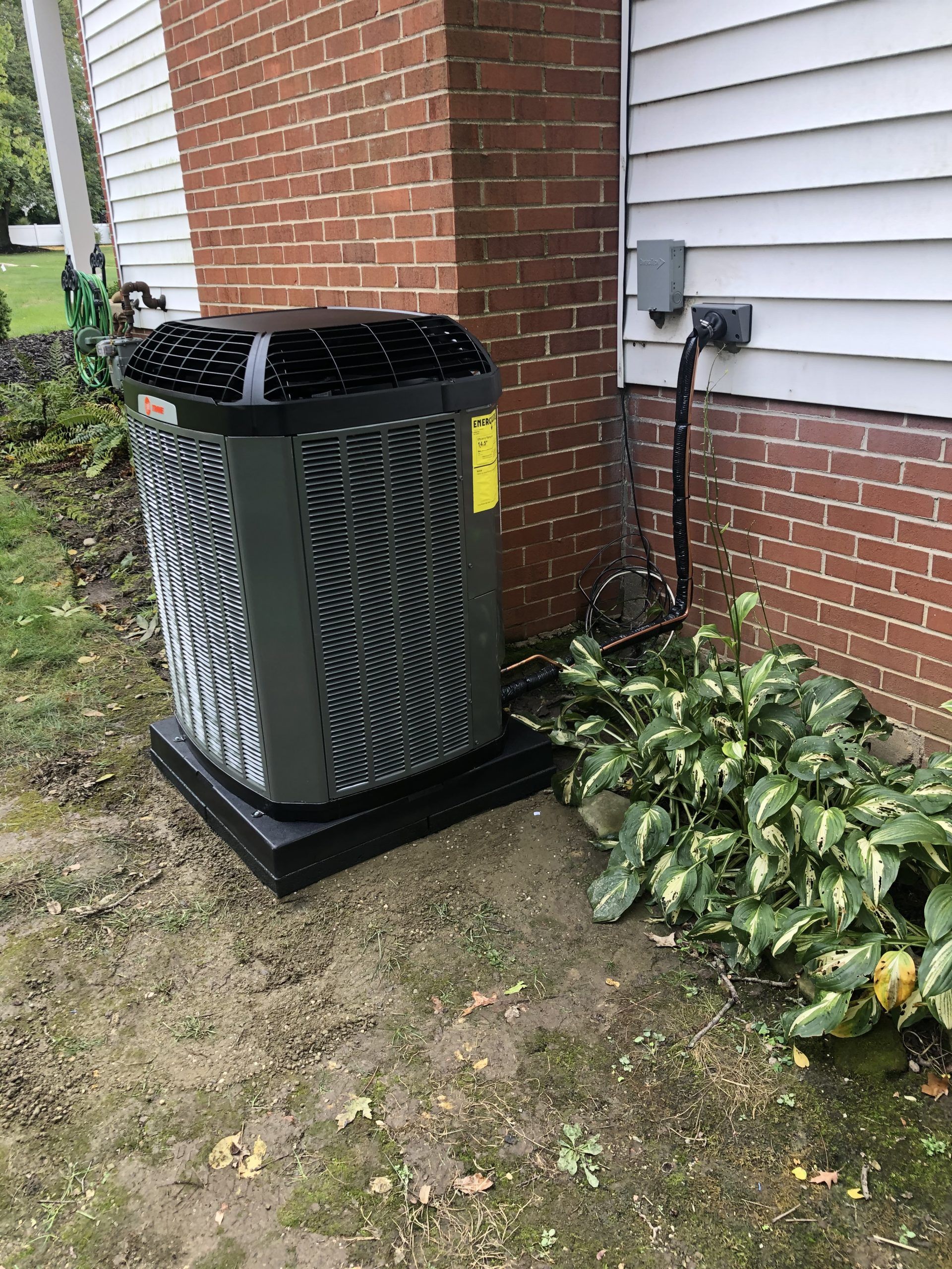 Outdoor air conditioning unit next to a brick wall and white siding, near greenery and electrical box.