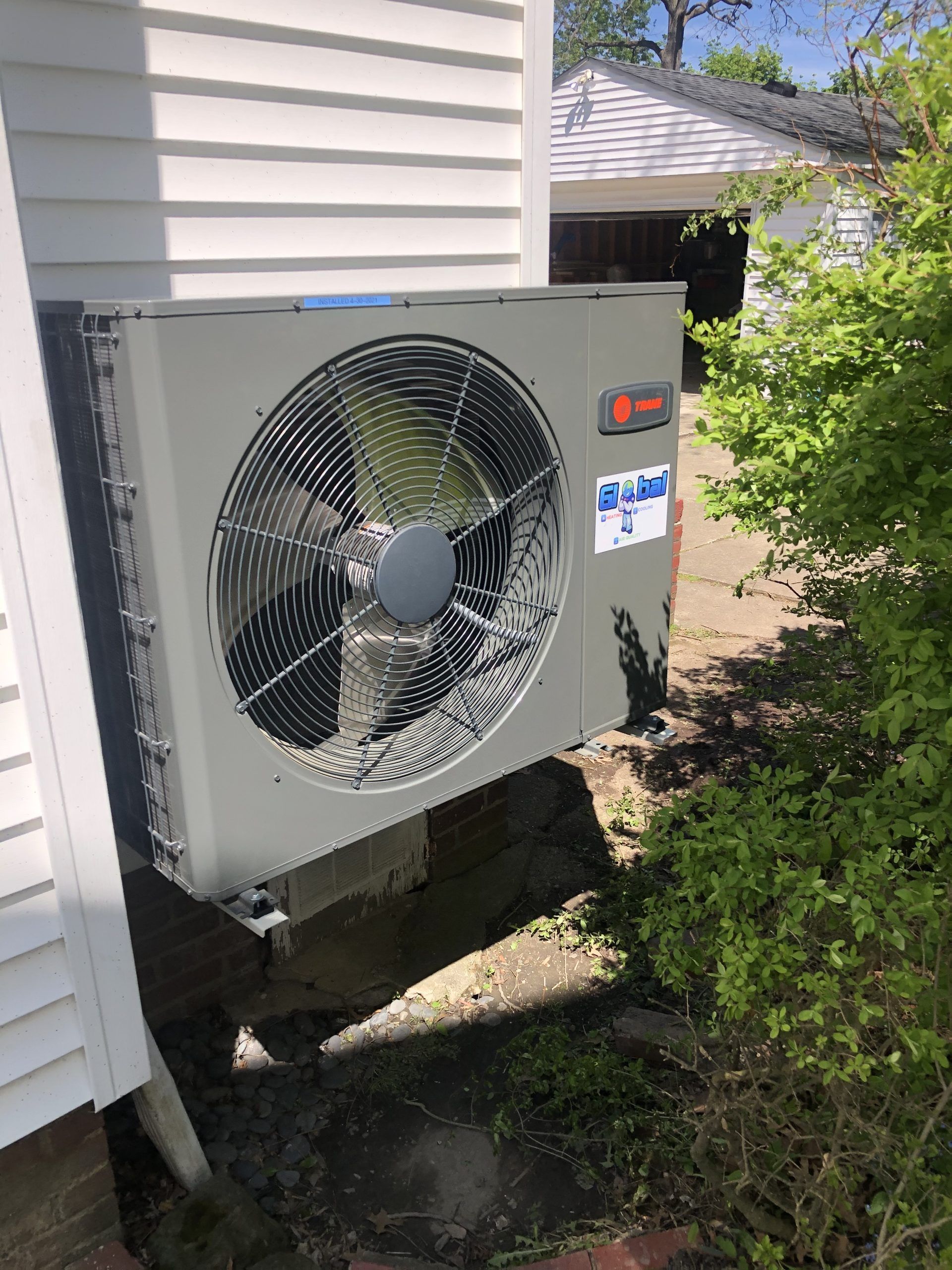 Trane heat pump unit mounted on a concrete block next to a white house with a garage in the background.