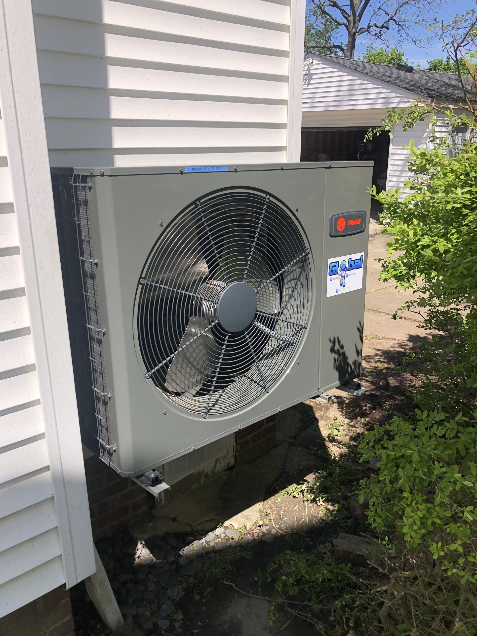 Air conditioner unit mounted on a wooden base next to a white house with a partially visible green yard.