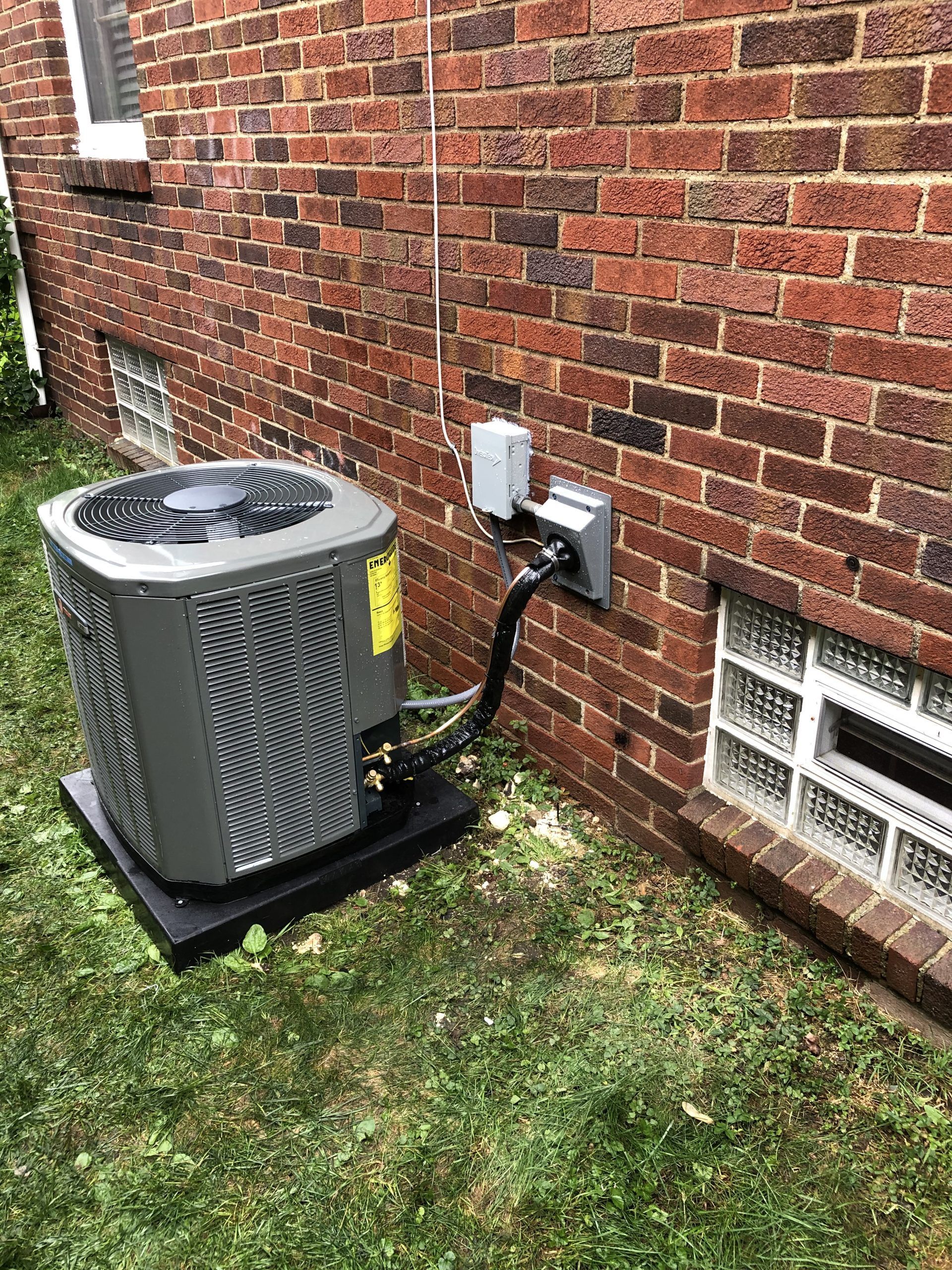 Air conditioner unit near a brick wall with electrical connections and small window.