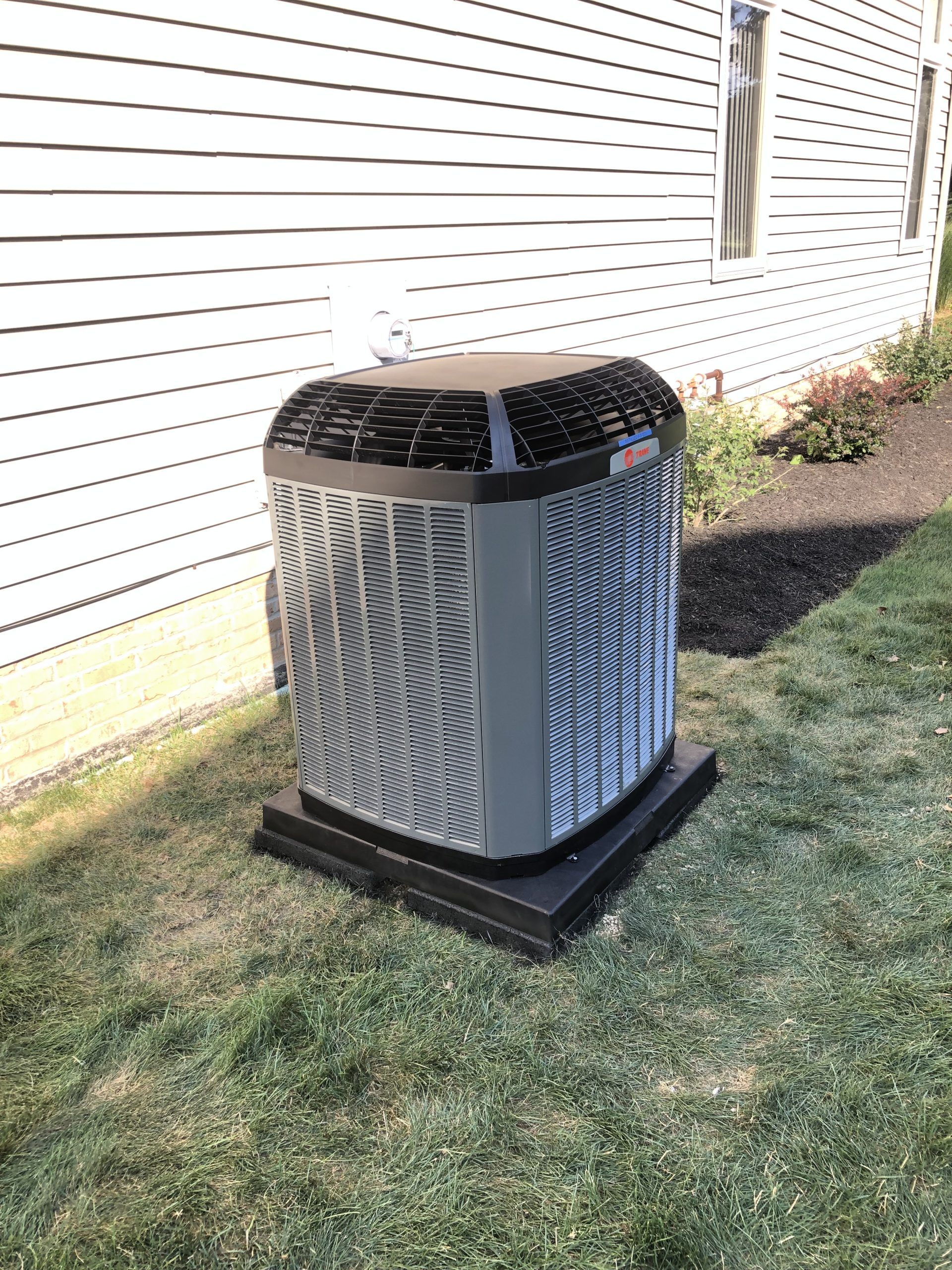 Air conditioning unit on a black base, next to a white house with landscaping.