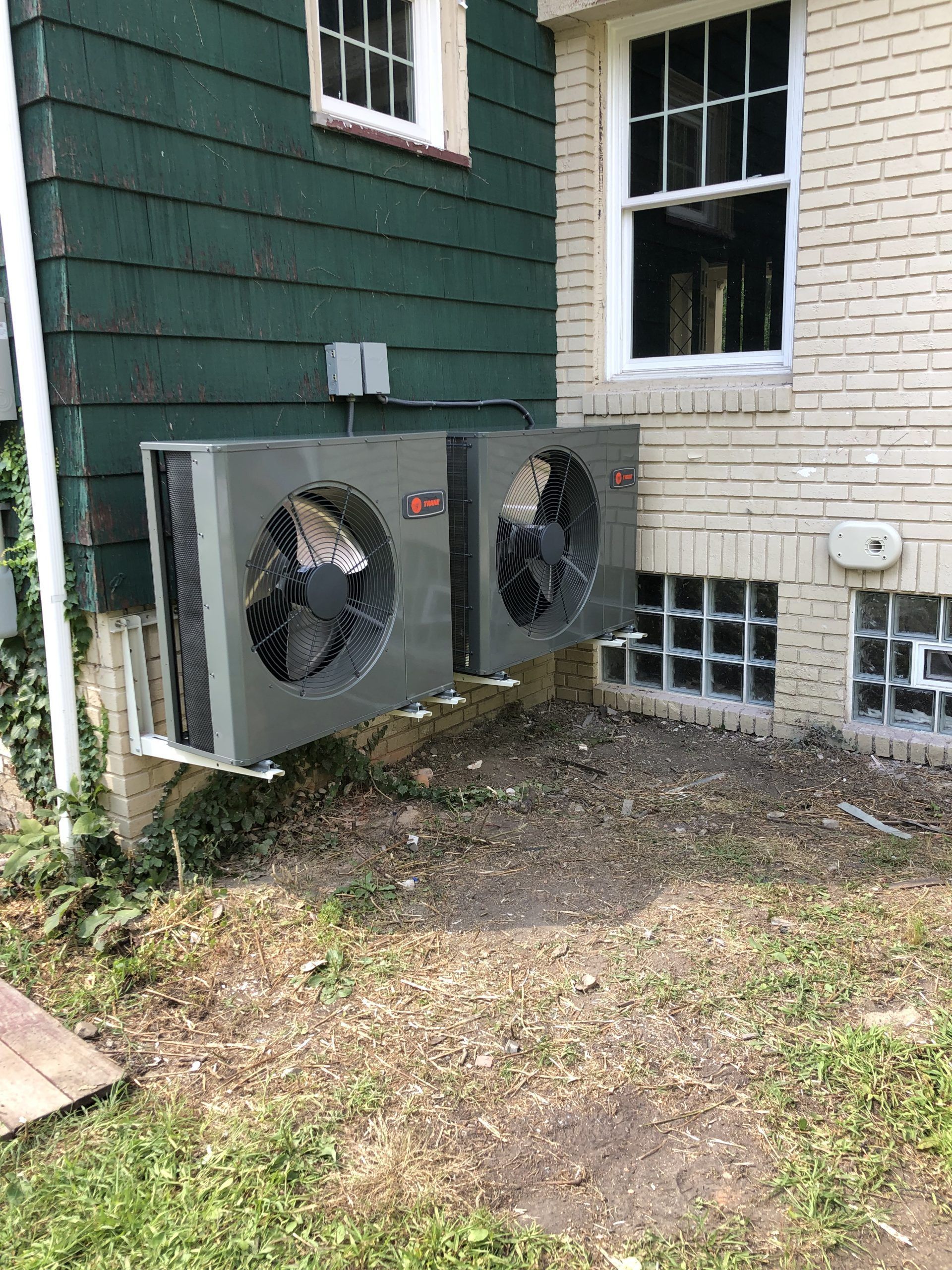 Two air conditioning units mounted on a building's exterior. One is on green siding, the other on brick.