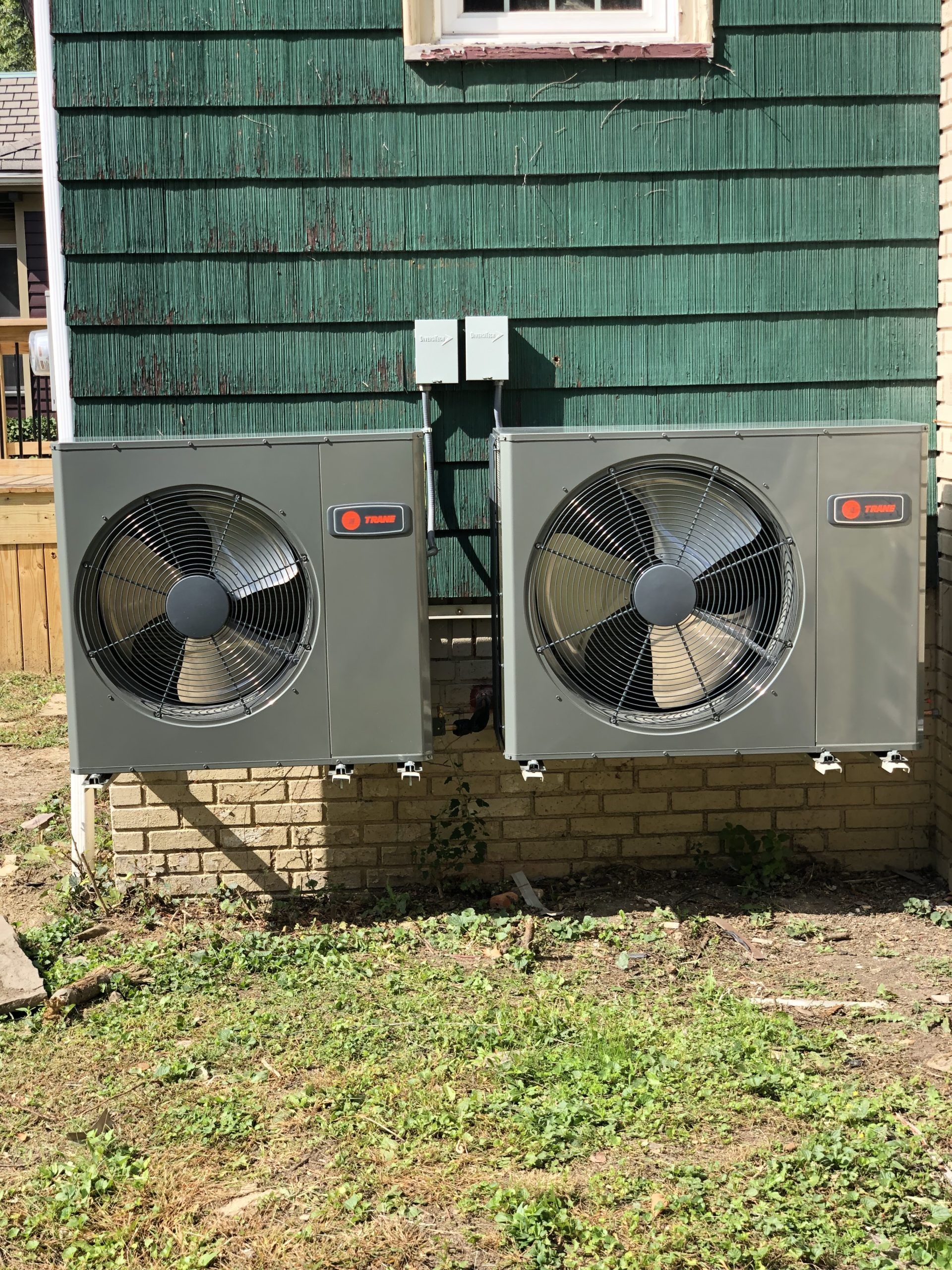 Two Trane heat pump units mounted on a brick wall with green siding. A small metal box is above them.