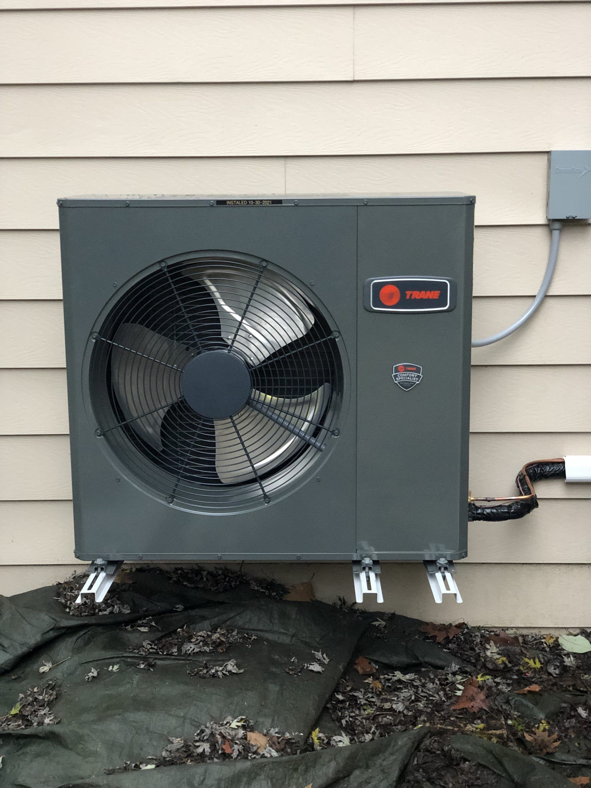 Gray Trane heat pump mounted on a beige wall, with a black fan and connecting pipes.